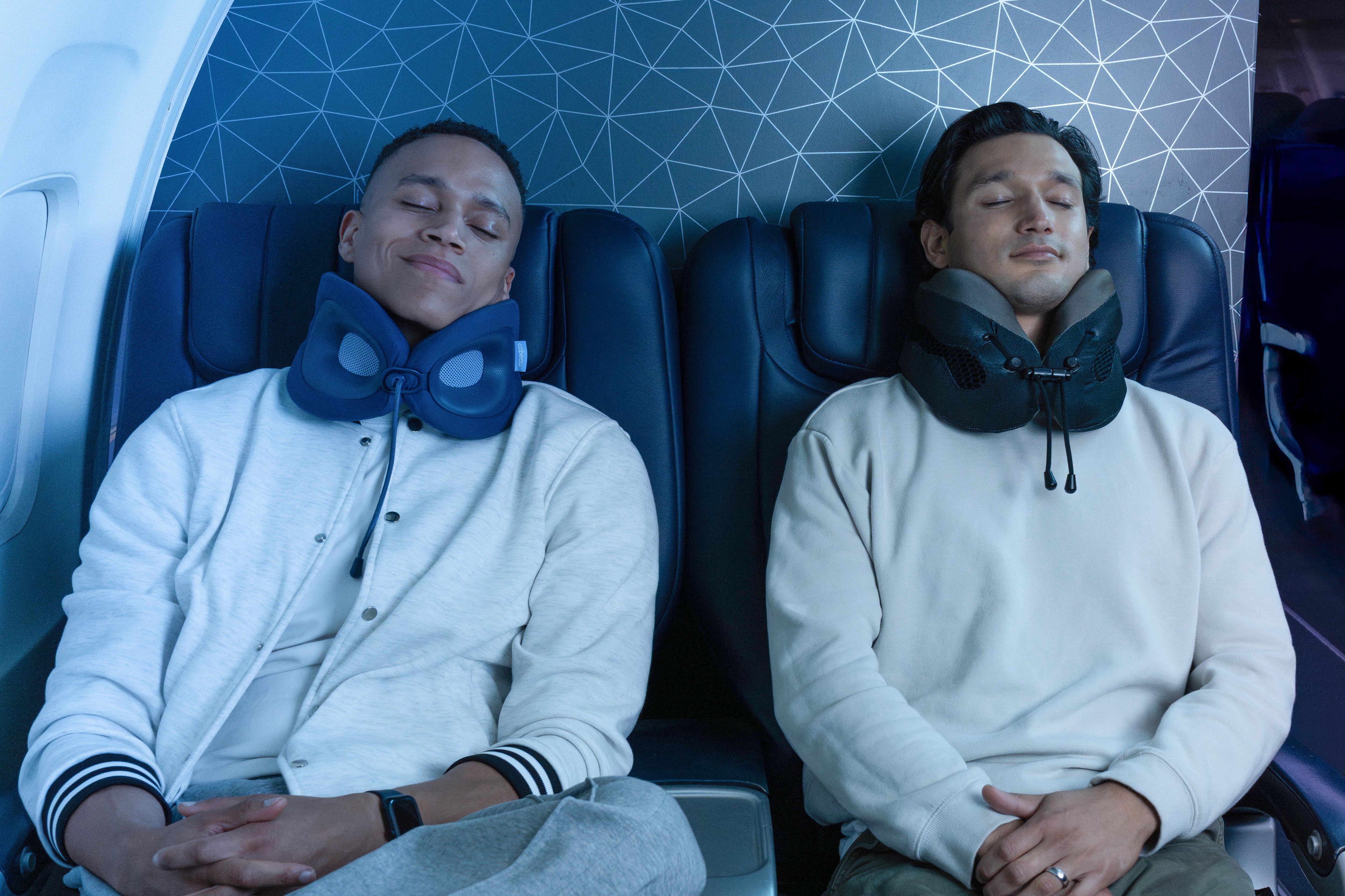 Two men sitting in airplane seats with neck pillows, one wearing a blue pillow and the other a green pillow.